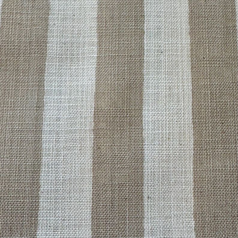 May include: A woven fabric featuring a repeating pattern of vertical stripes in alternating shades of light beige and off-white. The material displays a distinct, coarse, and textured weave characteristic of natural fibers, creating a uniform surface.
