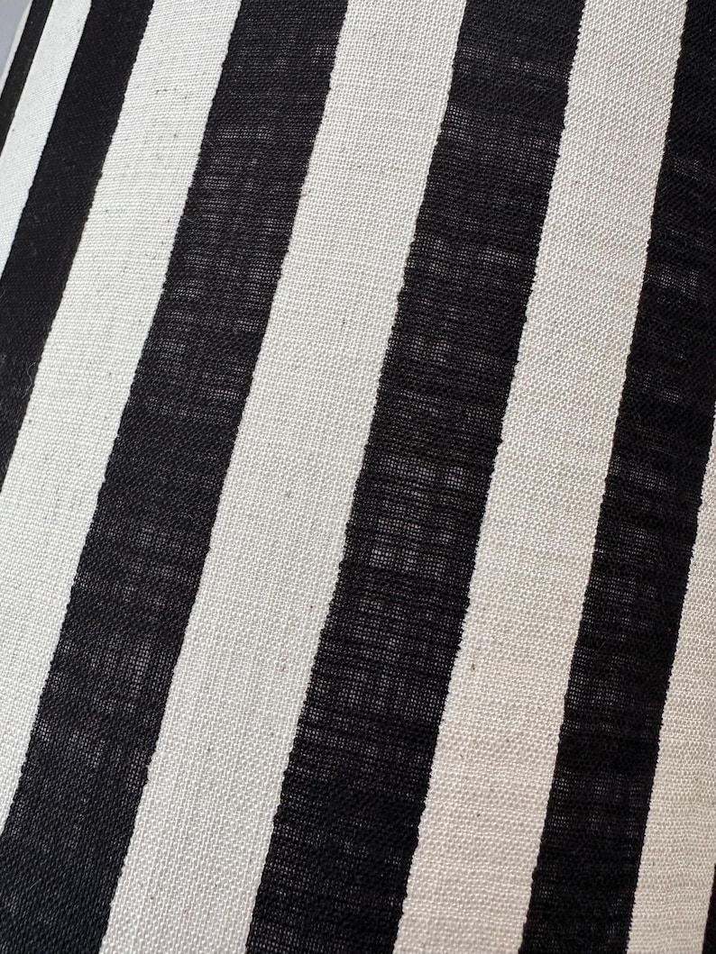 May include: A textile fabric featuring a repeating pattern of vertical stripes in alternating black and off-white colors. The material has a visible woven texture with a consistent weave throughout, creating a high-contrast linear design.