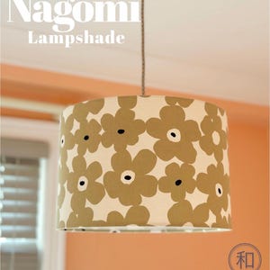 May include: A cylindrical lampshade featuring a repeating pattern of large, olive-green flowers with dark centers on a cream background. The text Nagomi Lampshade is visible at the top, and a circular logo with the Japanese character 和 is at the bottom.
