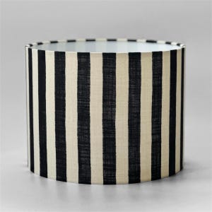 May include: A cylindrical drum lampshade featuring a repeating pattern of vertical black stripes set against a cream-colored fabric background. The material displays a visible woven texture throughout the surface of the shade, creating a consistent contrast between the dark stripes and the lighter base color.