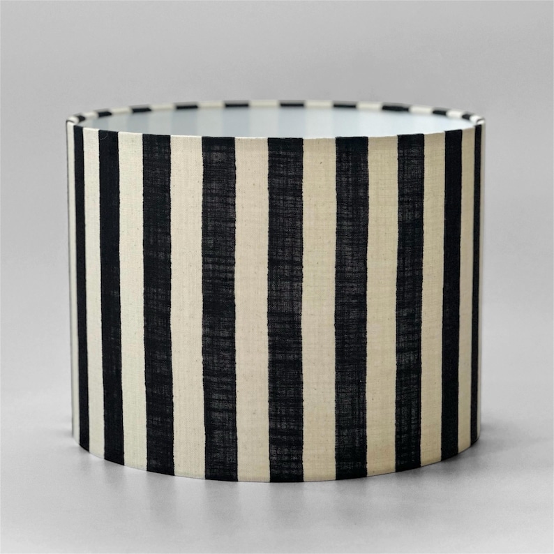 May include: A cylindrical drum lampshade featuring a repeating pattern of vertical black stripes set against a cream-colored fabric background. The material displays a visible woven texture throughout the surface of the shade, creating a consistent contrast between the dark stripes and the lighter base color.
