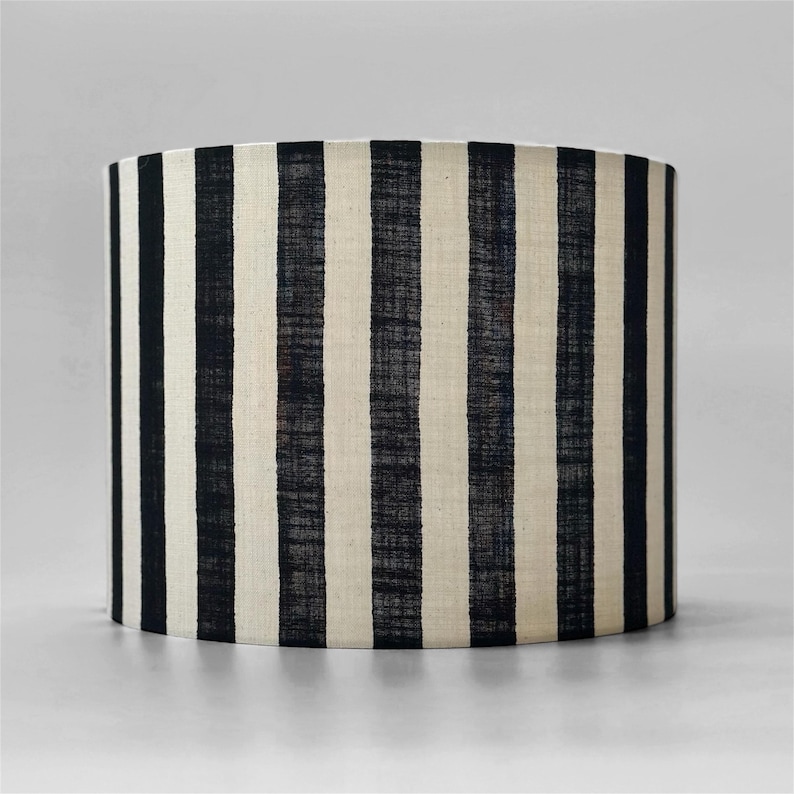 May include: A cylindrical lampshade featuring a repeating vertical stripe pattern in black and off-white. The fabric has a visible woven texture, and the design consists of alternating dark black stripes and lighter cream-colored stripes.