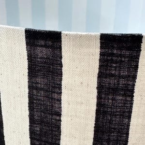 May include: A cylindrical lampshade featuring a bold vertical striped pattern in black and off-white. The fabric has a visible woven texture, and the interior of the shade displays a lighter, subtle blue and white vertical striped pattern.