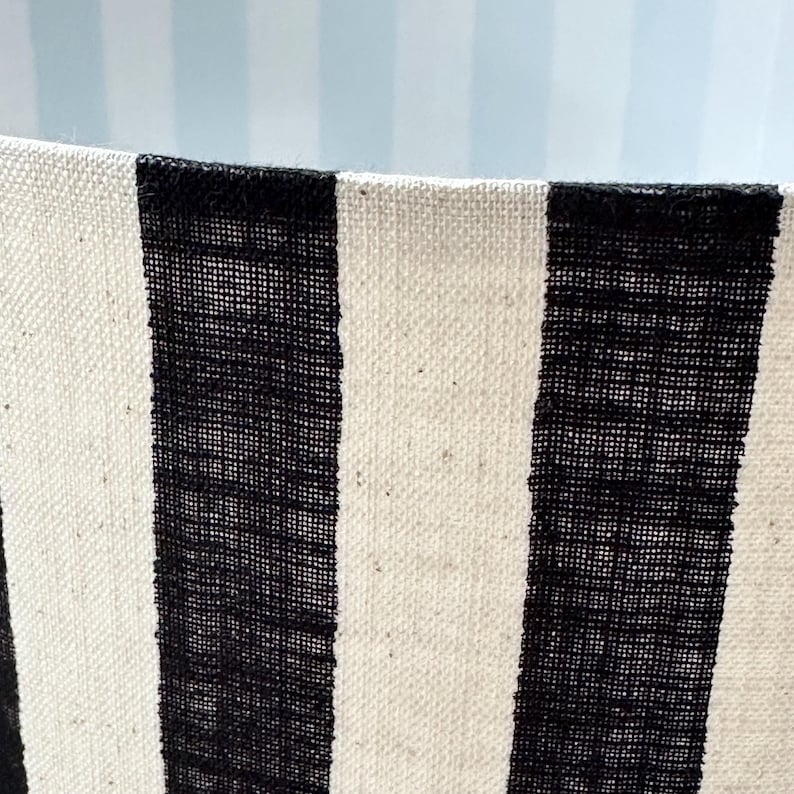 May include: A cylindrical lampshade featuring a bold vertical striped pattern in black and off-white. The fabric has a visible woven texture, and the interior of the shade displays a lighter, subtle blue and white vertical striped pattern.