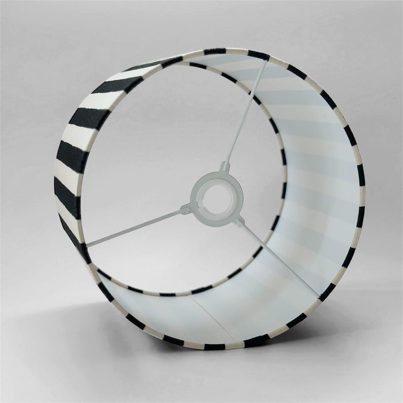 May include: A cylindrical lamp shade featuring a bold black and white striped pattern. The interior structure consists of a white metal frame with three spokes connecting to a central ring fitting, designed for attachment to a lamp base socket.