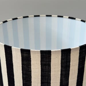May include: A cylindrical lamp shade featuring a bold black and white vertical striped exterior pattern. The interior lining of the lamp shade displays a subtle, light blue and white vertical striped design, visible from the top opening.