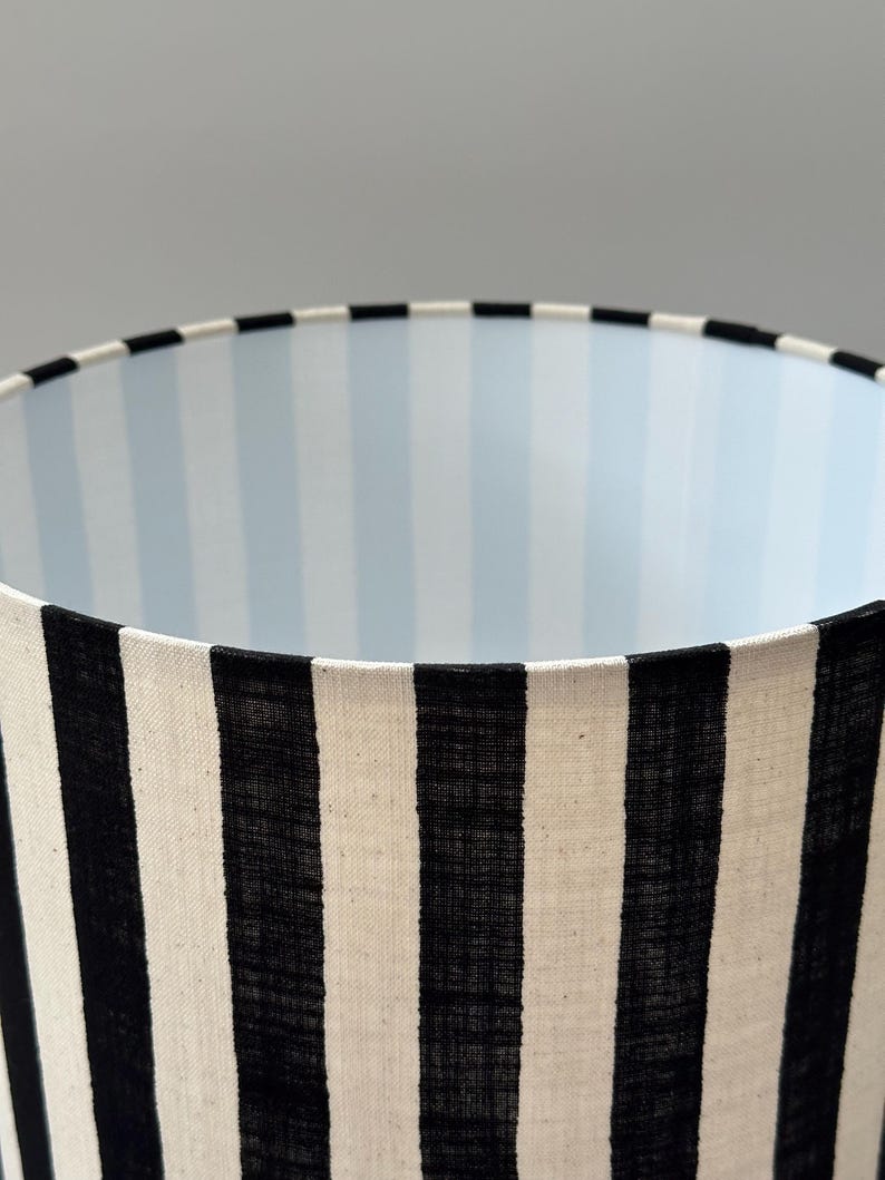 May include: A cylindrical lamp shade featuring a bold black and white vertical striped exterior pattern. The interior lining of the lamp shade displays a subtle, light blue and white vertical striped design, visible from the top opening.