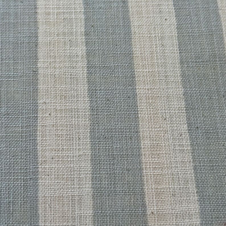 May include: A woven fabric featuring a repeating pattern of vertical stripes in alternating shades of muted slate blue and off-white. The textile displays a visible, textured grid-like weave structure throughout the entire surface of the material.