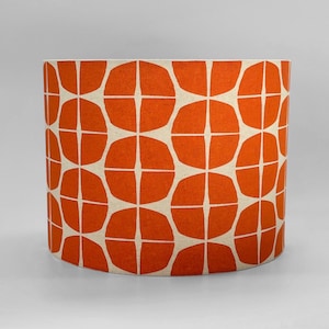 May include: A cylindrical drum lampshade featuring a repeating geometric pattern of bold orange circular shapes with central cross-like cutouts against a light cream-coloured fabric background, creating a consistent and symmetrical visual design.