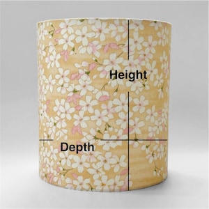 May include: A cylindrical lampshade featuring a light yellow background with a repeating pattern of white and pale pink cherry blossom flowers. Black lines are overlaid on the surface with the text Height and Depth indicating dimensions.