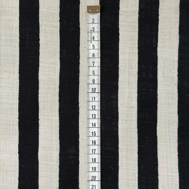May include: A fabric featuring a pattern of vertical black and off-white stripes. A white measuring tape is placed vertically over the fabric, showing markings from 2 to 21 inches, with the text Made in Germany printed near the top edge.