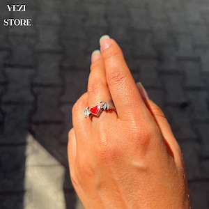 May include: A silver ring featuring a red heart and bat wings. The ring is worn on a person's finger.
