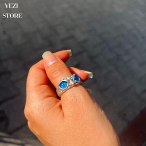 May include: Silver ring with two blue evil eye charms. The ring is worn on a finger.