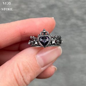 May include: A silver ring featuring a purple heart-shaped stone and a crown design. The ring is set with smaller clear stones.