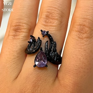 Puede incluir: Anillo negro con una piedra preciosa de color púrpura en forma de lágrima y un diseño de dos alas con una estrella en la parte superior.