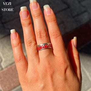 Può includere: Un anello d'argento con un designo a fiamma in smalto rosso. L'anello è indossato su un dito.