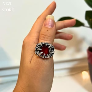 May include: A silver ring with a red gemstone set in a claw design. The ring is worn on a person's thumb.