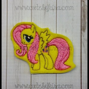 May include: Embroidered felt patch of Pinkie Pie from My Little Pony. The patch is yellow with pink hair and a blue eye. The patch is on a wood background.