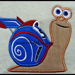 May include: Embroidered patch of a cartoon snail with a blue and red shell, wearing a racing helmet and goggles. The snail is carrying a number "5" on its shell. The text "www.cutebykira.com" is visible on the shell.