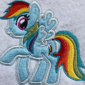 May include: Embroidered patch featuring a light blue pony with a rainbow-colored mane and tail. The pony has large eyes with pink and purple accents and white wings. The design is outlined with a silver thread.