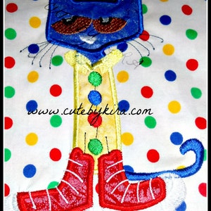 May include: Blue cat with brown eyes and a yellow shirt with green and blue buttons. The cat is wearing red boots and has a blue tail. The cat is on a white background with colorful polka dots.