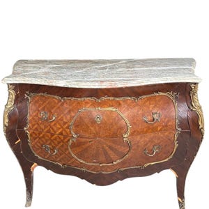 May include: An antique wooden chest of drawers with a marble top. The chest has a curved front with two drawers, ornate brass hardware, and decorative wood inlay. The legs are curved and the wood is a dark brown color.