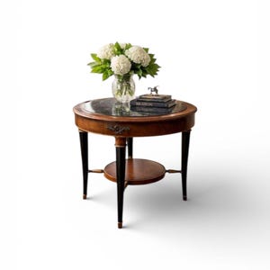 May include: A round side table with a dark marble top and a lower shelf. The table has a dark wood finish with gold accents and black legs. A vase of white flowers and books are on top.
