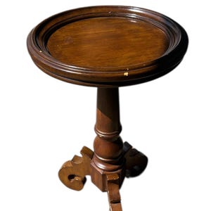 May include: A dark brown, antique-style side table. The table has a round top with a raised edge, supported by a turned pedestal and three decorative legs. The table is approximately 28 inches tall.