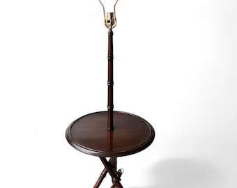May include: A dark brown bamboo-style floor lamp with a round table. The lamp has a brass-colored bulb holder and a dark brown wooden pole. The table is supported by three legs that cross at the bottom.