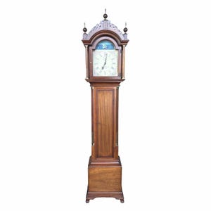 May include: A tall, antique grandfather clock made of dark wood. The clock features a detailed face with Roman numerals, a decorative top, and a long, slender body. The clock stands on a rectangular base and has a small door on the lower section.