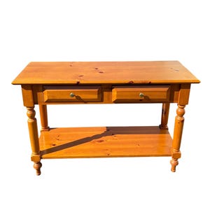 May include: A wooden console table with two drawers and a lower shelf. The table is made of light-colored wood and has turned legs. The drawers have brass knobs. The table is approximately 48 inches long, 16 inches wide, and 30 inches high.