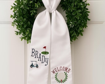 May include: White satin door sash with embroidered golf-themed designs. Features the name "Brady", a golf cart, a green with a red flag, and the word "WELCOME" with a golf ball and wreath.