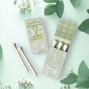 May include: Two rectangular cardboard matchbox-style containers with a light green and white tree pattern. One box is closed, displaying the text THANK YOU FOR SHOWERING, Est. 2020, and BABY VINCENT. The other box is open, revealing three white golf tees.