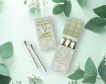 May include: Two rectangular cardboard matchbox-style containers with a light green and white tree pattern. One box is closed, displaying the text THANK YOU FOR SHOWERING, Est. 2020, and BABY VINCENT. The other box is open, revealing three white golf tees.