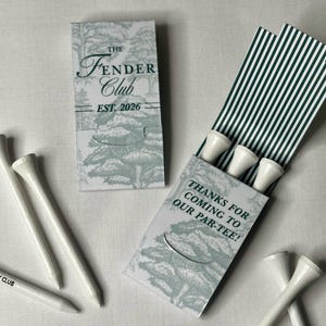 May include: A set of white golf tees and two rectangular paper matchbook-style holders. One holder features a green tree illustration and the text The Fender Club EST. 2026. The other holder displays the text THANKS FOR COMING TO OUR PAR-TEE!