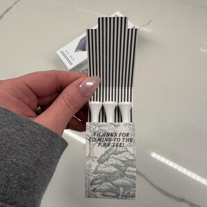 May include: A small rectangular cardboard box containing three white golf tees. The box features a black and white vertical striped interior flap and a gray and white forest illustration on the front with the text, THANKS FOR COMING TO THE PAR-TEE!