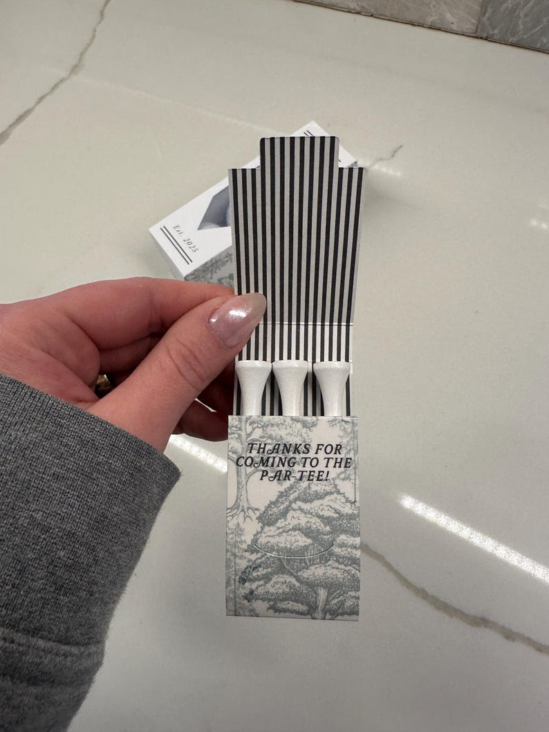 May include: A small rectangular cardboard box containing three white golf tees. The box features a black and white vertical striped interior flap and a gray and white forest illustration on the front with the text, THANKS FOR COMING TO THE PAR-TEE!