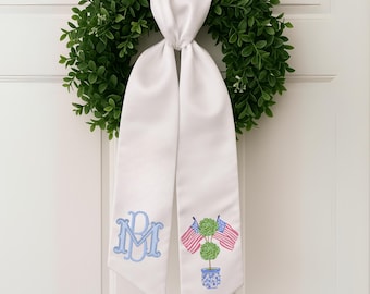 May include: White satin door hanger with a green wreath. The door hanger features a light blue monogram and an embroidered design of two American flags and topiary in a blue and white pot.
