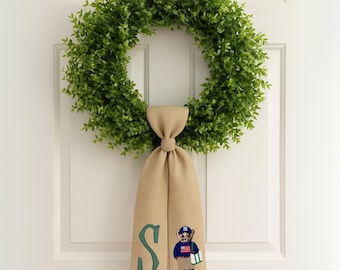 May include: A green, leafy wreath hangs on a white door. A tan fabric sash is tied to the wreath, featuring a green letter "S" and an embroidered bear wearing a baseball cap and holding a bag.