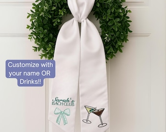May include: White satin sash with embroidered text and graphics. The sash is hanging on a green wreath. The text reads "Sarah's BACH CLUB" with a green bow graphic and two martini glasses. The text "Customize with your name OR Drinks!!" is also visible.