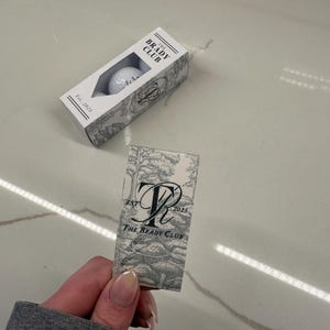 May include: A rectangular white box containing golf balls with a cutout window, labeled THE BRADY CLUB Est. 2025. Beside it is a matching rectangular card featuring a gray and white toile-style forest print, a large monogram, and the text THE BRADY CLUB EST 2025.