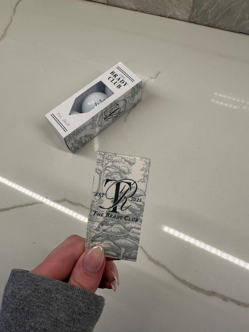May include: A rectangular white box containing golf balls with a cutout window, labeled THE BRADY CLUB Est. 2025. Beside it is a matching rectangular card featuring a gray and white toile-style forest print, a large monogram, and the text THE BRADY CLUB EST 2025.