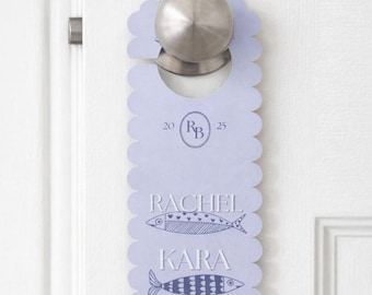 May include: A light purple door hanger with a scalloped edge, hanging on a white door. The text "RACHEL" and "KARA" are printed on the hanger, along with two fish illustrations and the initials "RB" inside a circle.