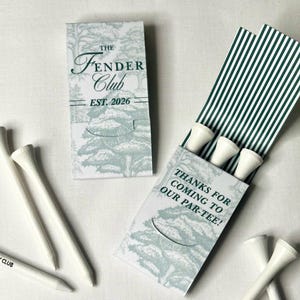 May include: Two rectangular matchbook-style paper sleeves featuring a light green forest illustration and dark green text. One sleeve reads The Fender Club Est. 2026, the other reads Thanks for coming to our par-tee. White golf tees and a pencil are included.