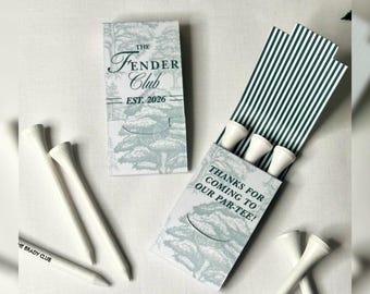 May include: Two rectangular matchbook-style paper sleeves featuring a light green forest illustration and dark green text. One sleeve reads The Fender Club Est. 2026, the other reads Thanks for coming to our par-tee. White golf tees and a pencil are included.
