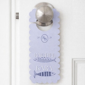 May include: A light blue, scalloped-edge door hanger template with the text "EDITABLE DOOR HANGER TEMPLATE." The design includes the names "RACHEL" and "KARA" with fish illustrations. The hanger is on a white door.