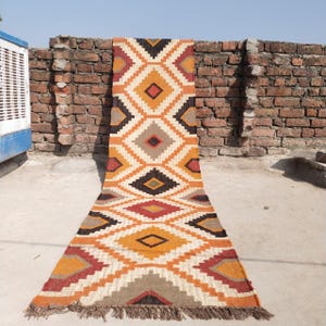 May include: A long, rectangular rug with a geometric diamond pattern in shades of orange, brown, cream, and black. The rug has a fringed edge and is displayed on a concrete surface against a brick wall.