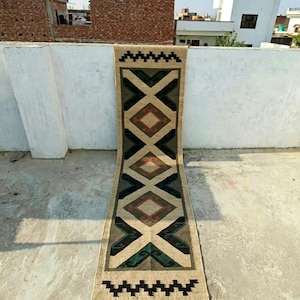 Puede incluir: Una alfombra pasillera larga y rectangular con base color canela y un patrón geométrico repetitivo. El diseño incluye triángulos de color verde oscuro y negro, rombos centrales en rojo apagado, y un borde en zigzag con flecos en el extremo.