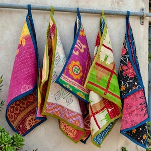Könnte beinhalten: Fünf rechteckige Stofftücher mit Kantha-Stickerei hängen an einer Metallstange. Jedes Tuch weist unterschiedliche Muster und Farben auf, darunter Magenta, Gelb, Grün und Schwarz, mit Blumen-, geometrischen und Gittermotiven sowie blauem Rand.