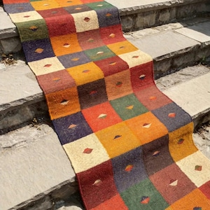 Puede incluir: Una alfombra de pasillo larga y rectangular con un patrón de cuadrícula repetitivo de cuadrados en varios colores, incluyendo rojo, naranja, amarillo, verde y azul oscuro. Cada cuadrado individual contiene un pequeño acento en forma de rombo centrado.